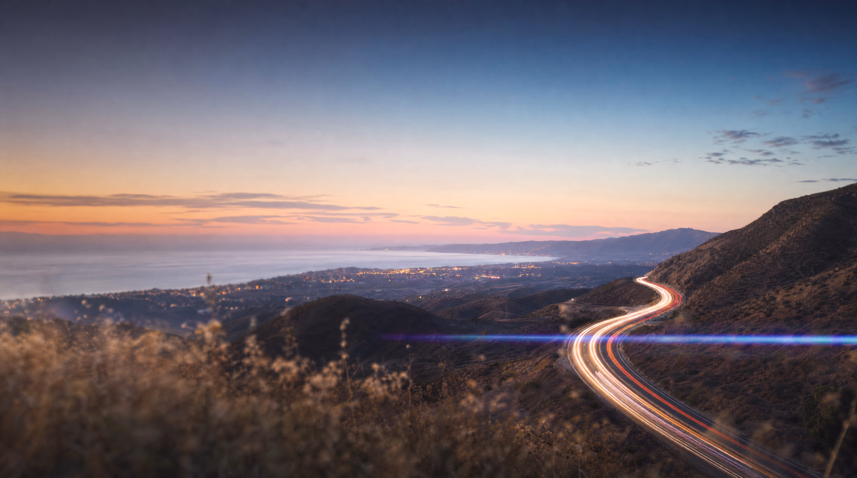 Coastal highway at dusk with long-exposure light trails winding through the hills toward the sea